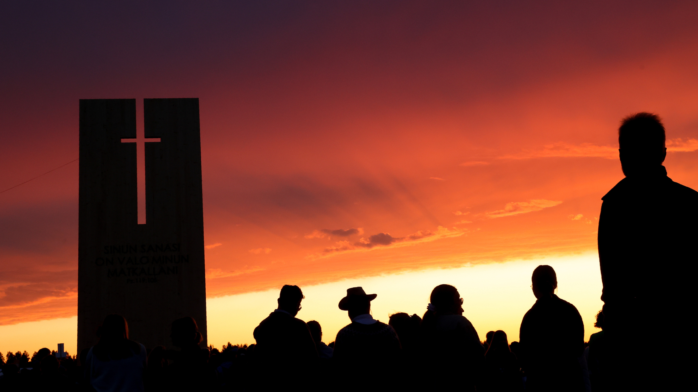 People are standing outside in a beautiful sunset, next to a festive port with a cross in it.